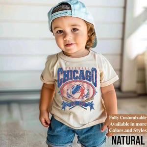 May include: A child wearing a beige t-shirt with a distressed Chicago baseball graphic in blue and red. The shirt has a crew neck and short sleeves. The child is also wearing blue denim shorts and a light blue baseball cap.