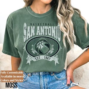 Może przedstawiać: Zielona koszulka z nadrukiem w stylu vintage z napisem "Basketball San Antonio" i wzorem koszyk&oacute;wki. Koszulka zawiera tekst "EST. 1967" i słowo "MOSS" na dole. Koszulka jest w stylu casual.