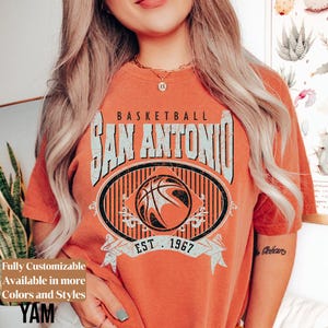 Może przedstawiać: Pomarańczowa koszulka z grafiką koszyk&oacute;wki. Projekt zawiera napis "BASKETBALL SAN ANTONIO" nad obrazem koszyk&oacute;wki, z napisem "EST. 1967" poniżej. Koszulka ma r&oacute;wnież napis "Fully Customizable Available in more Colors and Styles".