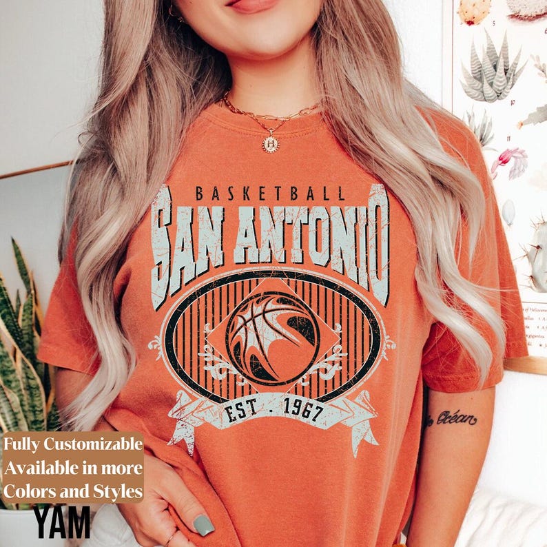 Może przedstawiać: Pomarańczowa koszulka z grafiką koszyk&oacute;wki. Projekt zawiera napis "BASKETBALL SAN ANTONIO" nad obrazem koszyk&oacute;wki, z napisem "EST. 1967" poniżej. Koszulka ma r&oacute;wnież napis "Fully Customizable Available in more Colors and Styles".