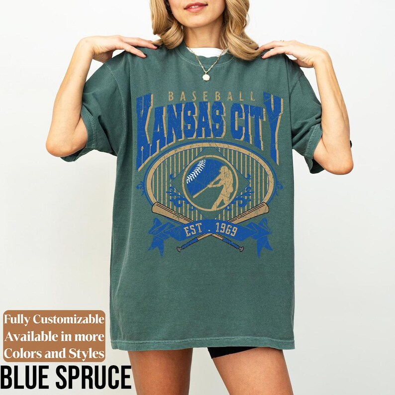 May include: Blue spruce-colored t-shirt with a baseball-themed graphic. The graphic features the text "Kansas City" in blue, along with a baseball, a batter silhouette, and crossed bats. The text "EST. 1969" is also included.