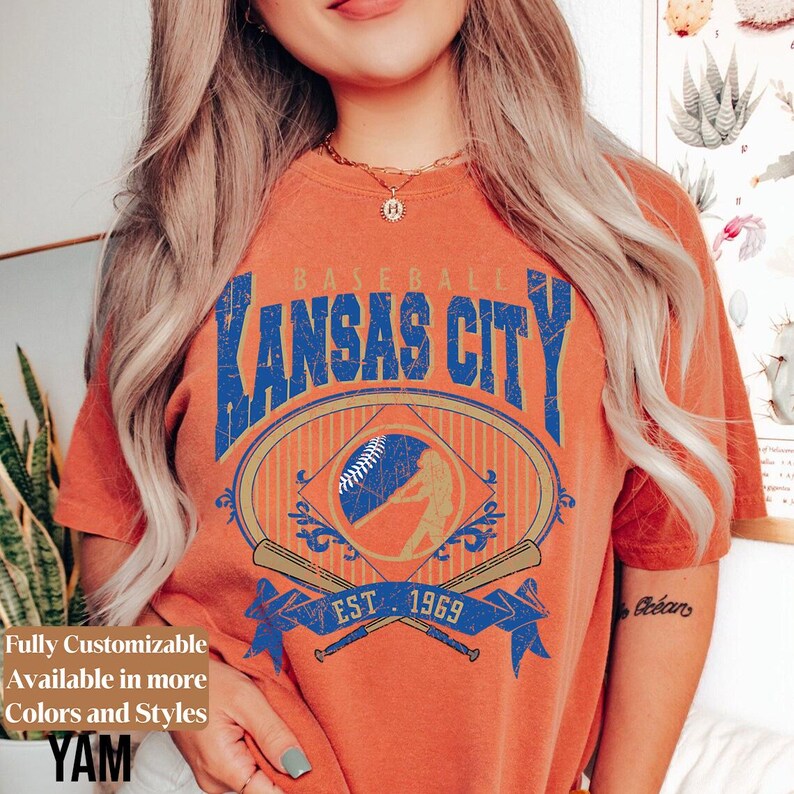 May include: Orange t-shirt with a distressed graphic design featuring "Kansas City" in blue lettering, a baseball, and crossed bats. The design includes the text "EST. 1969". The shirt also has the text "Fully Customizable Available in more Colors and Styles".