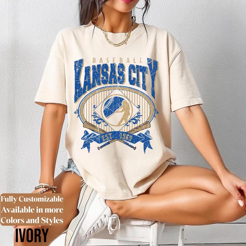 May include: Ivory-colored t-shirt featuring a distressed blue and gold baseball graphic that reads "Kansas City" with "Est. 1969". The shirt has short sleeves and a relaxed fit. The text "Fully Customizable Available in more Colors and Styles" is also visible.