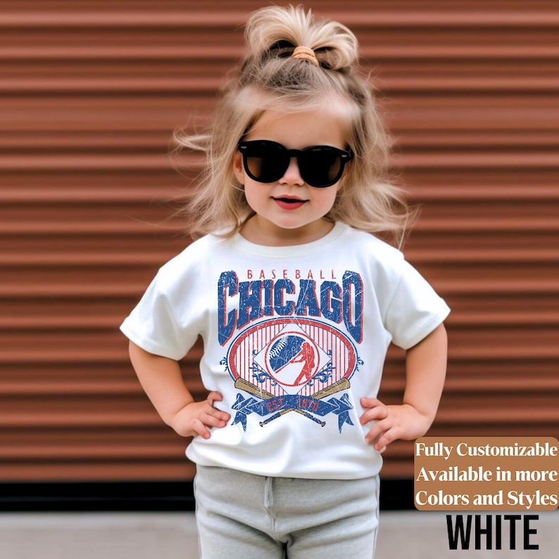 May include: A white t-shirt featuring a baseball-themed graphic with the text "CHICAGO BASEBALL" in blue and red. The design includes a baseball, bats, and the text "EST. 1971". The shirt is worn by a young child.