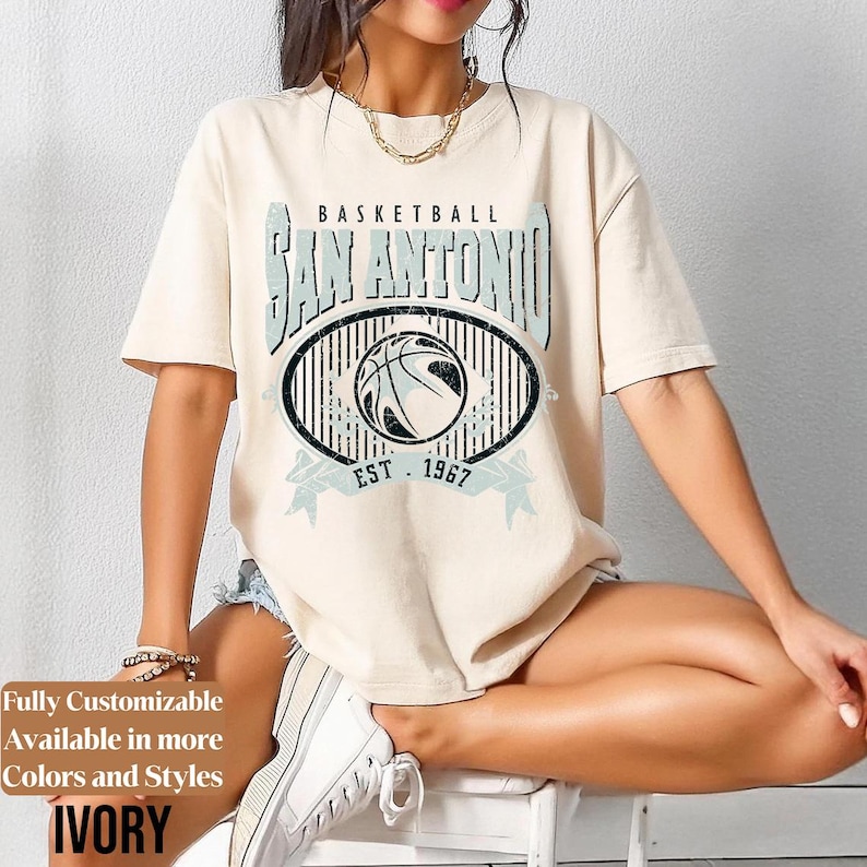 Może przedstawiać: Kremowa koszulka z grafiką koszyk&oacute;wki. Projekt zawiera napis "BASKETBALL SAN ANTONIO" nad owalem z koszyk&oacute;wką i napisem "EST. 1967". Koszulka jest w stylu casual.