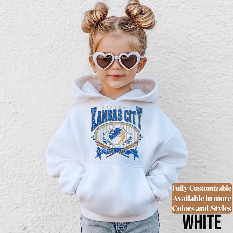 May include: A white hoodie with a Kansas City baseball graphic in blue and gold. The hoodie has a kangaroo pocket and a drawstring hood. The text "Fully Customizable Available in more Colors and Styles WHITE" is also visible.