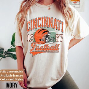 May include: An ivory t-shirt featuring a graphic print with the text CINCINNATI 1967 and Football in orange and black. The design includes an orange football helmet with black stripes and a face mask, set against a faded green background.
