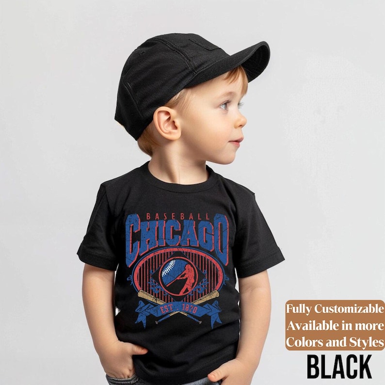 May include: A black t-shirt featuring a baseball-themed graphic with the word "Chicago" in blue and red. The design includes a baseball, a batter, and crossed bats. The shirt is worn with a black cap. The text "EST. 1870" is also visible.
