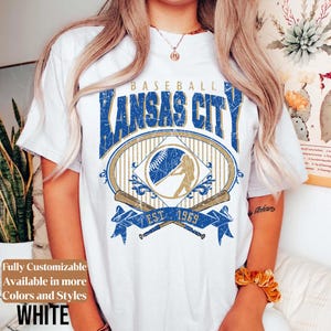 May include: White t-shirt featuring a distressed blue and gold graphic with the text "BASEBALL KANSAS CITY EST. 1969". The design includes a baseball, a batter, and crossed bats. The shirt is available in more colors and styles.