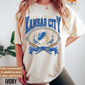 May include: Ivory-colored t-shirt featuring a distressed blue and gold graphic that reads "Baseball Kansas City" with a baseball and player silhouette. The shirt also has the text "EST. 1969".