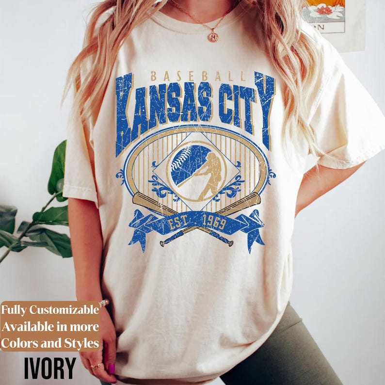 May include: Ivory-colored t-shirt featuring a distressed blue and gold graphic that reads "Baseball Kansas City" with a baseball and player silhouette. The shirt also has the text "EST. 1969".