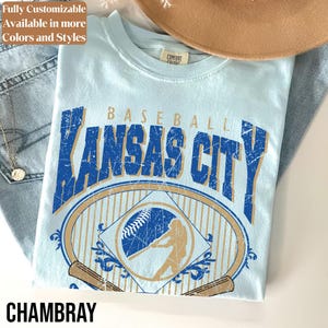 May include: Light blue t-shirt with a distressed graphic that says "Baseball Kansas City". The design includes a baseball, a batter silhouette, and baseball bats. The word "Chambray" is at the bottom.