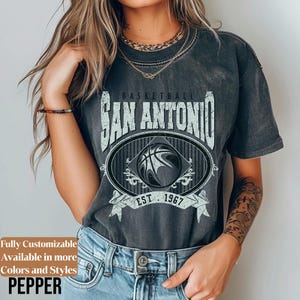 Może przedstawiać: Ciemnoszara koszulka z efektem postarzanego nadruku. Projekt zawiera napis "BASKETBALL SAN ANTONIO" nad owalnym emblematem z koszyk&oacute;wką i napisem "EST. 1967". Słowo "PEPPER" znajduje się na dole.