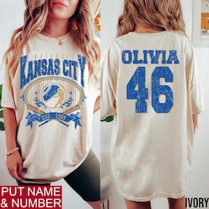 May include: Ivory-colored baseball t-shirt with "Kansas City" in blue lettering and a baseball graphic on the front. The back displays "Olivia" and the number "46" in blue. The shirt has a vintage, distressed look.