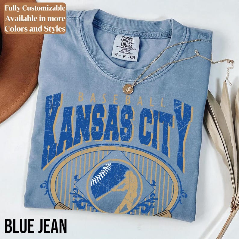 May include: A blue jean colored t-shirt with a distressed graphic that says "Kansas City Baseball" in blue and gold. The shirt has a crew neck and short sleeves. A gold necklace with a pendant rests on the shirt.