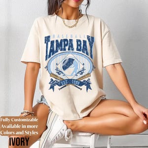 May include: Ivory-colored baseball t-shirt featuring a Tampa Bay design. The design includes the words "Baseball Tampa Bay" in blue, along with a baseball and a batter silhouette. The shirt also has "EST. 1998" on a banner. The shirt is a relaxed fit.