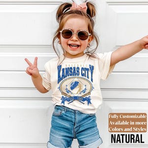 May include: A young child wearing a cream-colored t-shirt with a blue and gold "Kansas City Baseball" graphic. The child is also wearing denim shorts and sunglasses. The shirt has the text "EST. 1969".