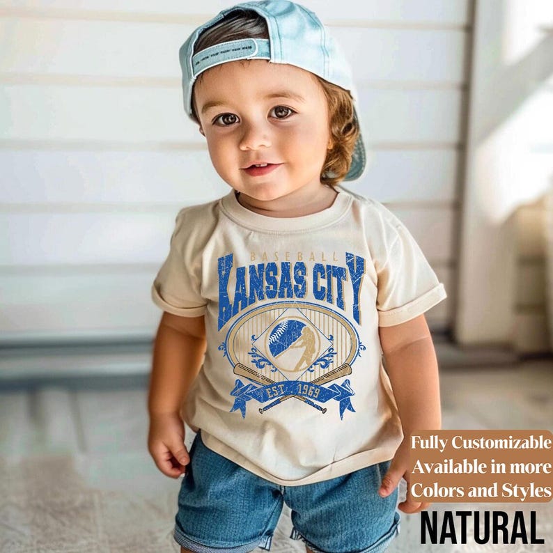 May include: A child wearing a beige t-shirt and a light blue baseball cap. The shirt features a blue graphic with "Kansas City Baseball" and a baseball design. The text "EST. 1969" is also visible. The child is also wearing blue denim shorts.