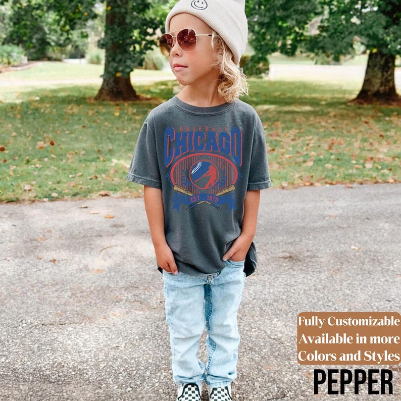 May include: A child wearing a charcoal gray t-shirt with a vintage-style Chicago baseball design in red and blue. The child is also wearing light blue jeans, a beige beanie, and sunglasses. The text "Fully Customizable Available in more Colors and Styles PEPPER" is visible.