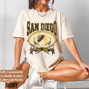 May include: Ivory-colored t-shirt with a distressed "San Diego Baseball" graphic. The design includes a baseball, a batter silhouette, crossed bats, and the year "1969". The shirt has short sleeves and a relaxed fit.