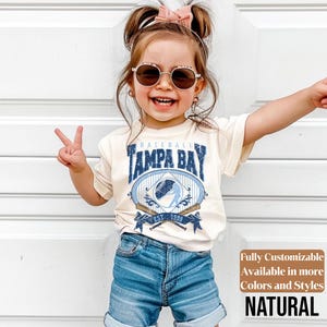 May include: A young child wearing a cream-colored t-shirt with a "Tampa Bay Baseball" design in blue and white. The shirt features a baseball graphic and the text "EST. 1998". The child is also wearing denim shorts and sunglasses.