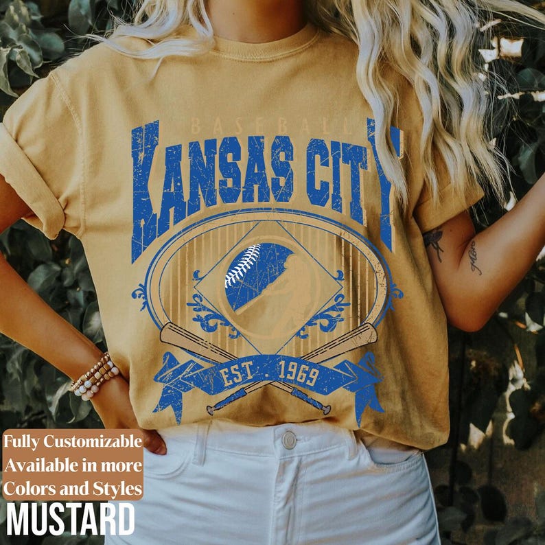 May include: Mustard-colored t-shirt with a distressed blue graphic featuring "Kansas City" above a baseball design. The design includes a baseball, crossed bats, and the text "EST. 1969". The shirt is part of a customizable collection.