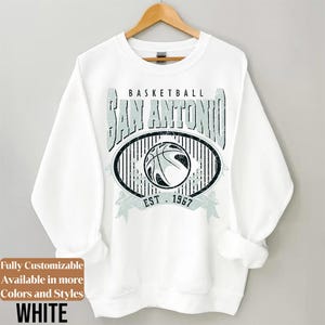 Może przedstawiać: Biała bluza z nadrukiem koszyk&oacute;wki. Projekt zawiera napis "BASKETBALL SAN ANTONIO EST. 1967" w stylu vintage, z koszyk&oacute;wką pośrodku. Bluza wisi na drewnianym wieszaku.