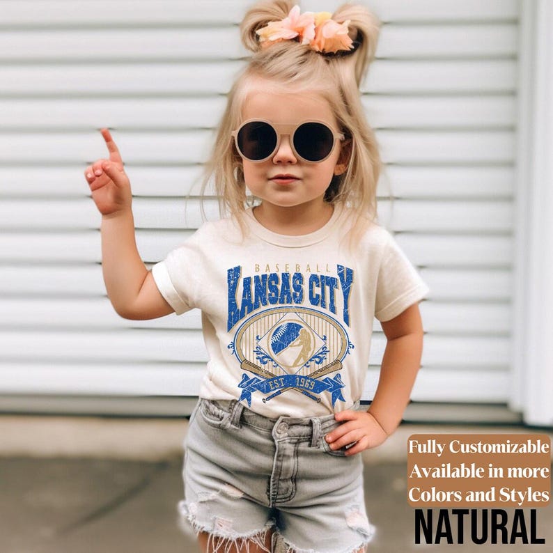 May include: A cream-colored t-shirt featuring a baseball-themed graphic with the text "Kansas City" in blue. The design includes a baseball, bats, and the year "EST. 1969". The shirt is paired with gray denim shorts.