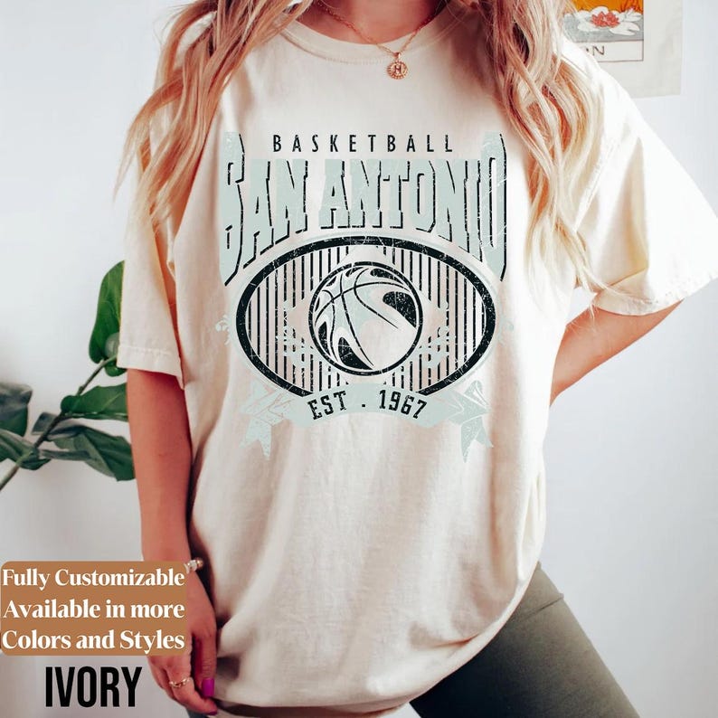 Może przedstawiać: Kremowa koszulka z motywem koszyk&oacute;wki. Projekt zawiera napis "BASKETBALL SAN ANTONIO" w postarzanym kroju pisma, grafikę koszyk&oacute;wki i rok "EST. 1967". Koszulka ma luźny kr&oacute;j.