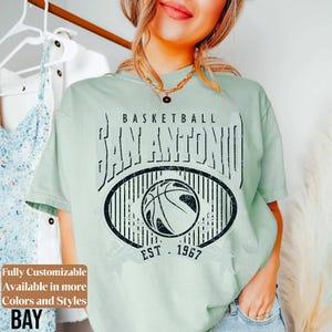 Może przedstawiać: Jasnozielona koszulka z czarnym nadrukiem graficznym. Projekt zawiera tekst "BASKETBALL SAN ANTONIO EST. 1967" i obraz koszyk&oacute;wki. Część kolekcji odzieżowej.