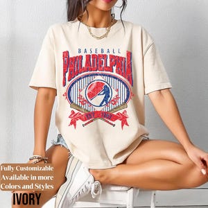 May include: Ivory-colored t-shirt with a distressed baseball graphic. The graphic features the word "BASEBALL" above "PHILADELPHIA" in red, with a baseball and batter illustration. The text "EST. 1883" is below the graphic.
