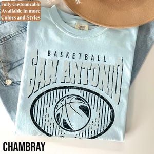 Może przedstawiać: Jasnoniebieska koszulka z nadrukiem "BASKETBALL SAN ANTONIO" nad wzorem koszyk&oacute;wki. Słowo "CHAMBRAY" jest wydrukowane na dole. Koszulka jest złożona na dżinsach i brązowym kapeluszu.