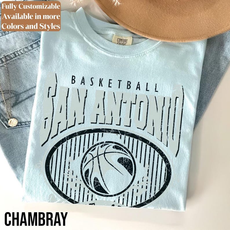 Może przedstawiać: Jasnoniebieska koszulka z nadrukiem "BASKETBALL SAN ANTONIO" nad wzorem koszyk&oacute;wki. Słowo "CHAMBRAY" jest wydrukowane na dole. Koszulka jest złożona na dżinsach i brązowym kapeluszu.