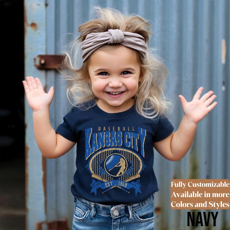 May include: A navy blue t-shirt with a baseball-themed graphic that says "Kansas City" in a vintage style. The shirt features a baseball and a batter, with the text "EST. 1969". The image also includes the text "Fully Customizable Available in more Colors and Styles NAVY".