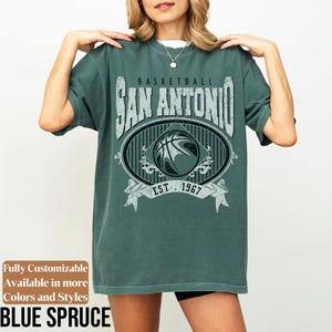 Może przedstawiać: Niebieska koszulka z nadrukiem w stylu vintage. Grafika zawiera napis "BASKETBALL SAN ANTONIO EST. 1967" wok&oacute;ł wzoru koszyk&oacute;wki. Koszulka ma luźny kr&oacute;j.