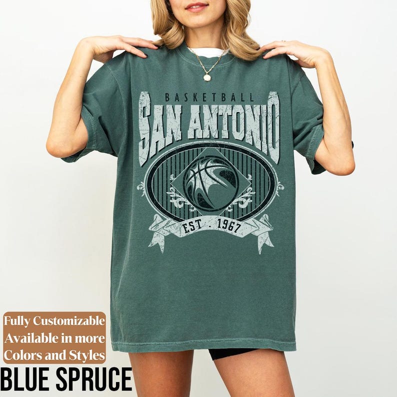 Może przedstawiać: Niebieska koszulka z nadrukiem w stylu vintage. Grafika zawiera napis "BASKETBALL SAN ANTONIO EST. 1967" wok&oacute;ł wzoru koszyk&oacute;wki. Koszulka ma luźny kr&oacute;j.