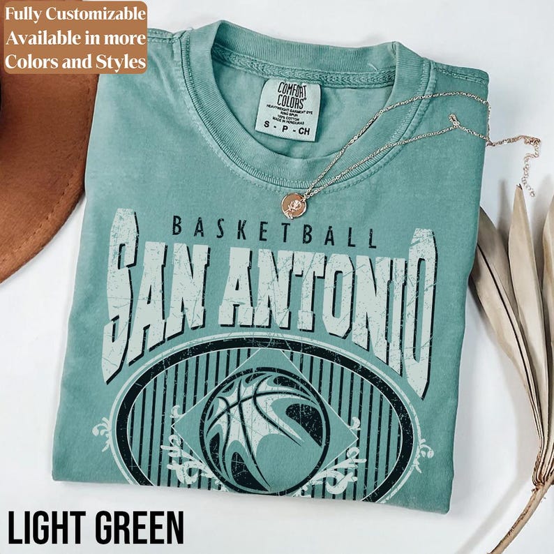 Może przedstawiać: Jasnozielona koszulka z postarzanym wzorem graficznym z napisem "BASKETBALL SAN ANTONIO" i obrazem koszyk&oacute;wki. Koszulka ma okrągły dekolt i kr&oacute;tkie rękawy. Srebrny naszyjnik z wisiorkiem spoczywa na koszulce. Tekst "LIGHT GREEN" znajduje się na dole.