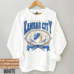 May include: White sweatshirt with a baseball-themed graphic. The design features the text "KANSAS CITY BASEBALL" in blue, along with a baseball, a batter silhouette, and crossed bats. The text "EST. 1969" is also included.
