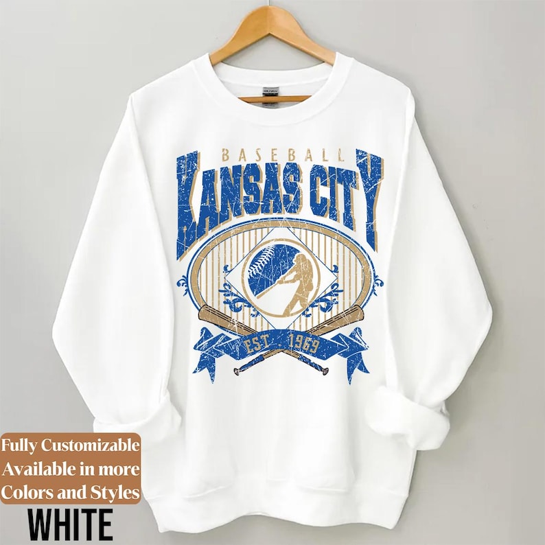 May include: White sweatshirt with a baseball-themed graphic. The design features the text "KANSAS CITY BASEBALL" in blue, along with a baseball, a batter silhouette, and crossed bats. The text "EST. 1969" is also included.