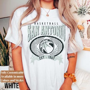 Może przedstawiać: Biała koszulka z szarą, postarzaną grafiką z napisem "BASKETBALL SAN ANTONIO EST. 1967" i motywem koszyk&oacute;wki. Koszulka ma r&oacute;wnież tekst "Fully Customizable Available in more Colors and Styles WHITE".