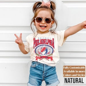 May include: A young child wearing a cream-colored t-shirt with a baseball-themed graphic that says "Philadelphia" in red and blue. The child is also wearing denim shorts and sunglasses. The text "Fully Customizable Available in more Colors and Styles NATURAL" is in the bottom right corner.