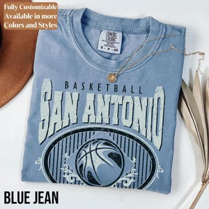 Może przedstawiać: Niebieska koszulka dżinsowa z nadrukiem "BASKETBALL SAN ANTONIO" i wzorem koszyk&oacute;wki. Koszulka ma okrągły dekolt i kr&oacute;tkie rękawy. Złoty naszyjnik z wisiorkiem jest zarzucony na koszulkę.