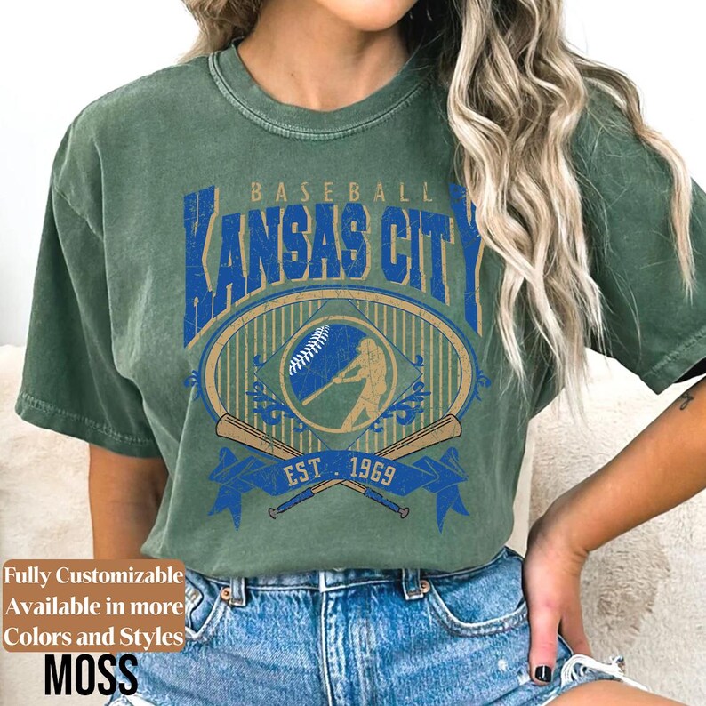 May include: Moss green t-shirt featuring a vintage baseball design. The design includes the text "Baseball Kansas City" in blue, a baseball graphic, crossed bats, and "EST. 1969". The shirt is part of a customizable collection.