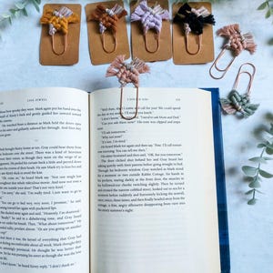May include: Five colorful macrame bookmarks with tassels, each attached to a copper paperclip. The bookmarks are displayed on a brown background with a book open in the background.