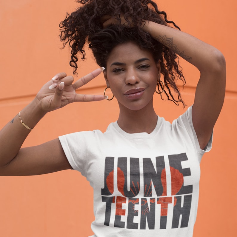 May include: A woman wearing a white t-shirt with the text "JUNE TEENTH" in black letters with a red and orange graphic design.