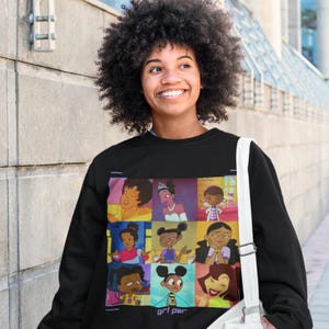 May include: Black sweatshirt with a colorful graphic print of cartoon characters. The graphic features nine different characters, including a princess, a doctor, and a bee. The text "girl pwr" is printed below the graphic.