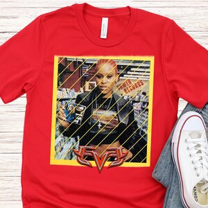 Op de afbeelding: Rood T-shirt met een afbeelding van rapper Eve. De afbeelding toont Eve voor een Tower Records-achtergrond, met het logo van de artiest onderaan. Witte Converse sneakers en een jeans zijn zichtbaar.