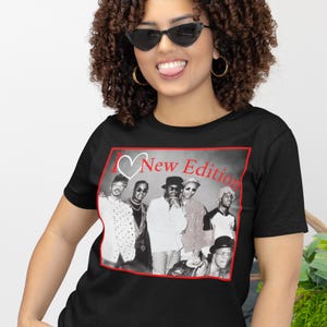 May include: Black t-shirt featuring a black and white image of a music group with the text "I ❤️ New Edition" in red. The image is framed in a red border. The shirt is worn by a person wearing sunglasses.
