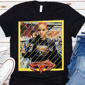 Op de afbeelding: Zwart T-shirt met een afbeelding van een vrouw die een cd vasthoudt in een geel omlijst beeld met diagonale gele lijnen. Het shirt heeft een rood en zilver "EVE" logo aan de onderkant.