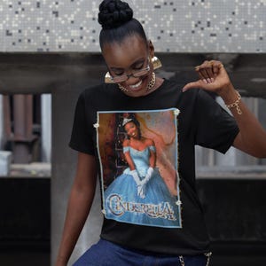 May include: Black t-shirt featuring a portrait of Cinderella in a blue gown, with the word "Cinderella" in a decorative font. The image has a vintage style with a light blue border and floral accents.